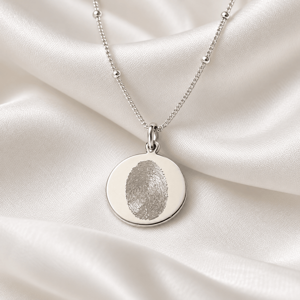 A silver necklace with a round pendant featuring an engraved fingerprint, displayed on draped white satin fabric.